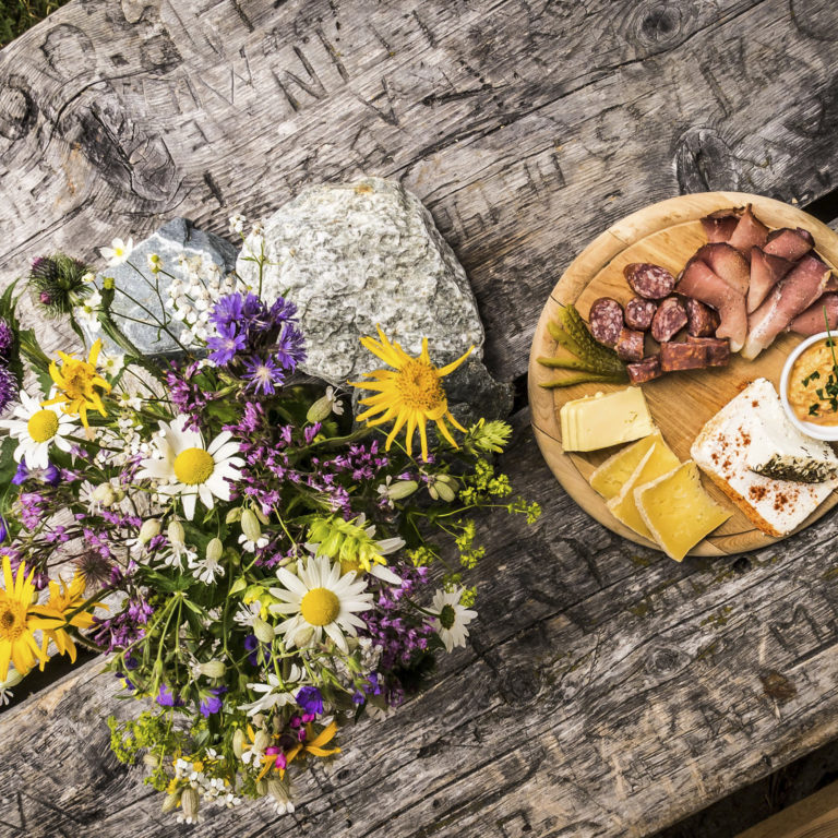 Brotzeit, eine Jause mit Speck und Käse auf der Alpe Garnera im Montafon, mit herbstlichem Blumenstrauß auf dem Tisch