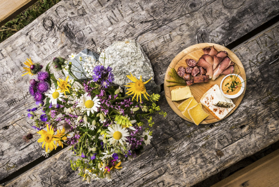 Brotzeit, eine Jause mit Speck und Käse auf der Alpe Garnera im Montafon, mit herbstlichem Blumenstrauß auf dem Tisch