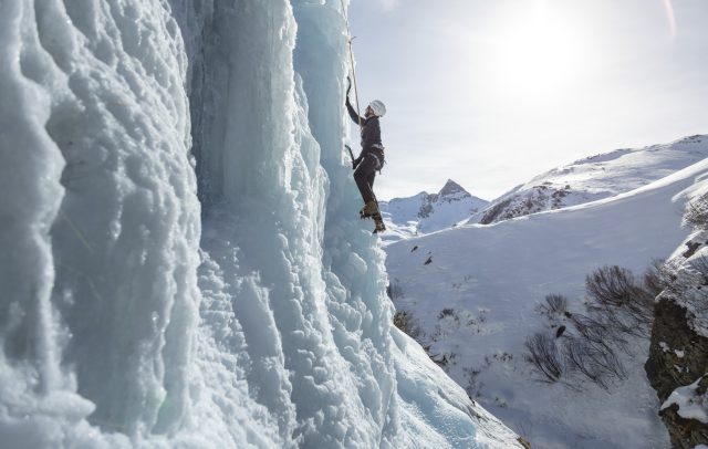 Ein Eiskletterer klettert mit Steigeisen und Pickel eine Eiswand hinauf, gesichert mit einem Seil.