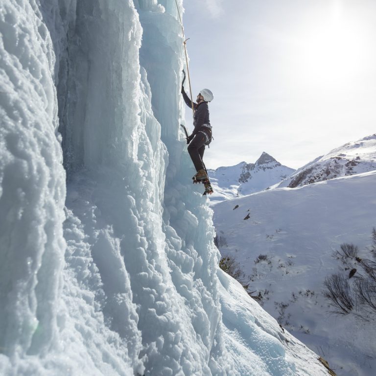 Ein Eiskletterer klettert mit Steigeisen und Pickel eine Eiswand hinauf, gesichert mit einem Seil.