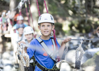 Kinder mit Kletterhelm und Klettergurt überqueren angeseilt eine Seilbrücke im Hochseilgarten in Gargellen im Montafon.