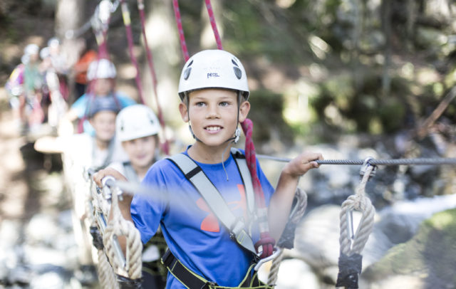 Kinder mit Kletterhelm und Klettergurt überqueren angeseilt eine Seilbrücke im Hochseilgarten in Gargellen im Montafon.
