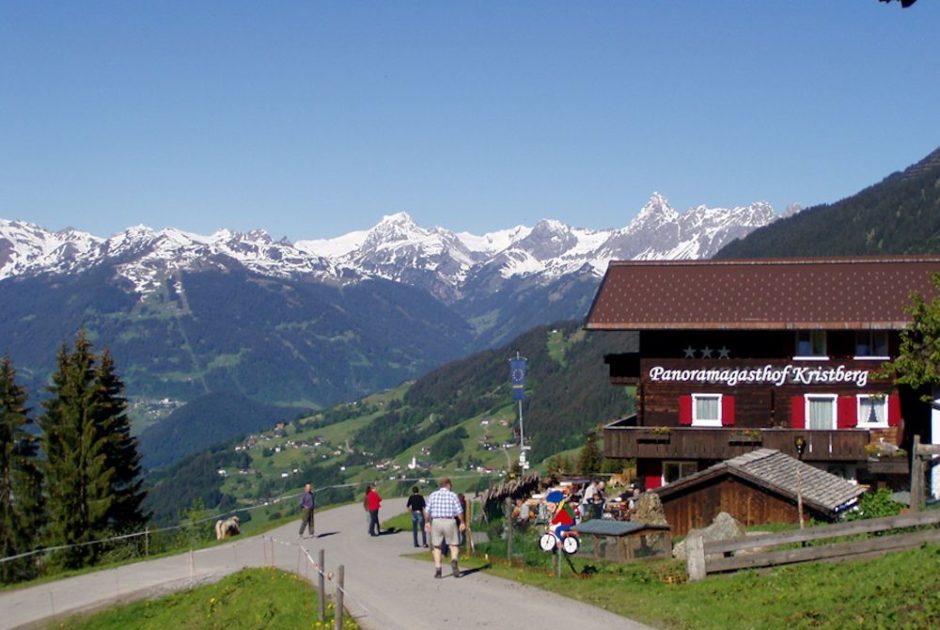 Der aus dunklem Holz gebaute Panoramagasthof Krsitberg mit grünen Wiesen. Im Hintergrund die Montafoner Bergwelt mit weißen Berggipfeln und saftig grünen Tälern.