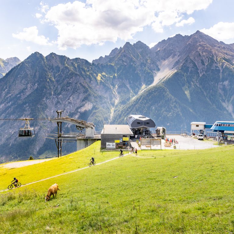 Die Bergstation der Bergbahnen Brandnertal mit eintreffender Gondel. Auf der Wiese davor grasen Kühe, im Hintergrund die Berge des Brandnertals