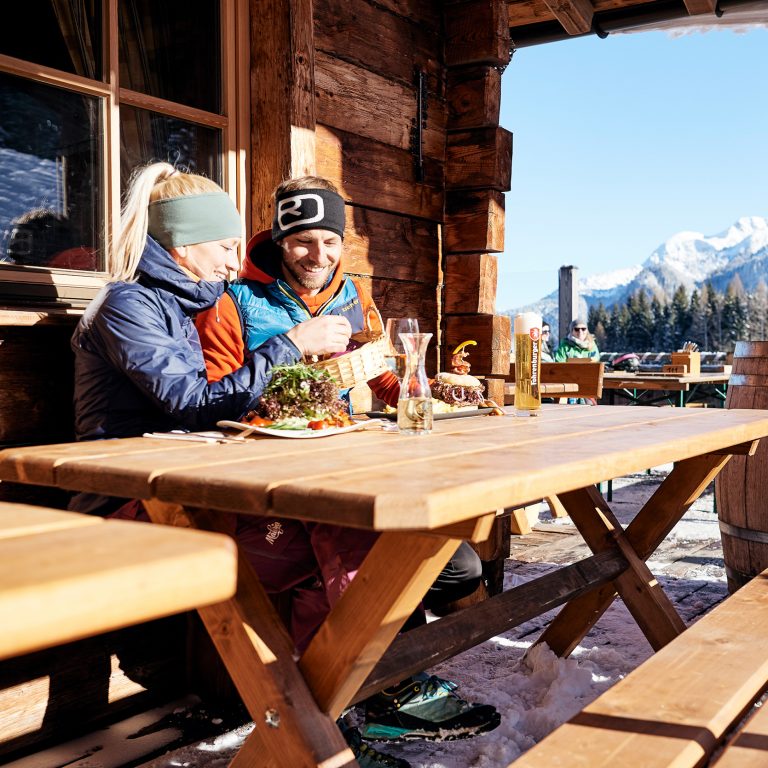 Eine Frau und ein Mann sitzen auf der Terrasse einer Berghütte, auf dem Tisch vor ihnen stehen Speisen und Getränke, im Hintergrund ein schneebedeckter Berg