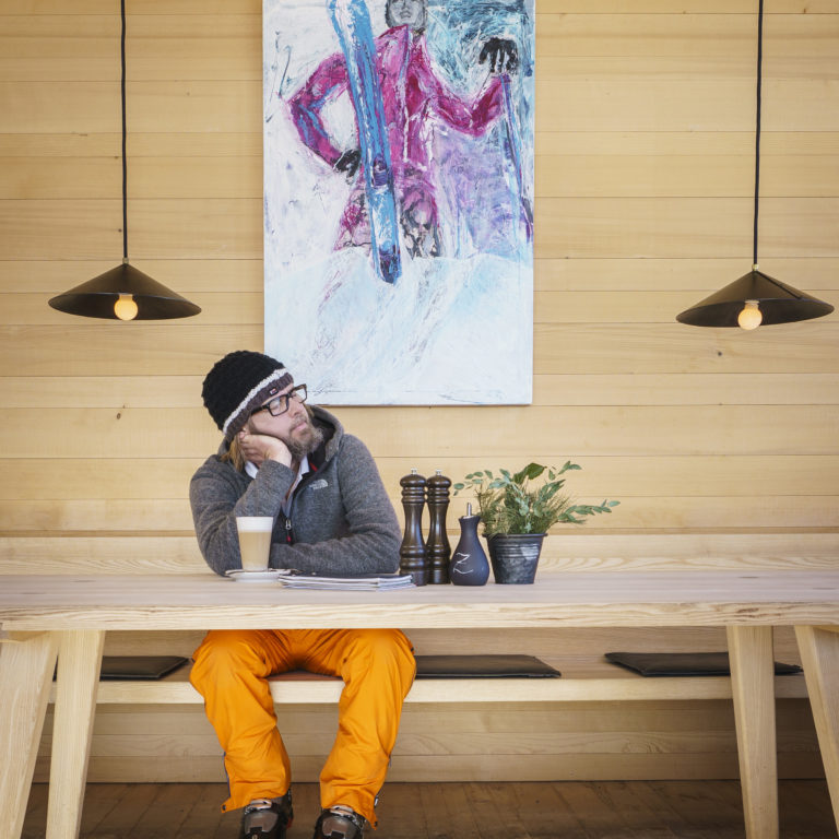 Ein Mann sitzt an einem Tisch, stützt mit dem Ellenbogen auf dem Tisch und seinen Kopf in der Hand, er hat die Augen geschlossen und vor ihm steht ein Kaffeegetränk.