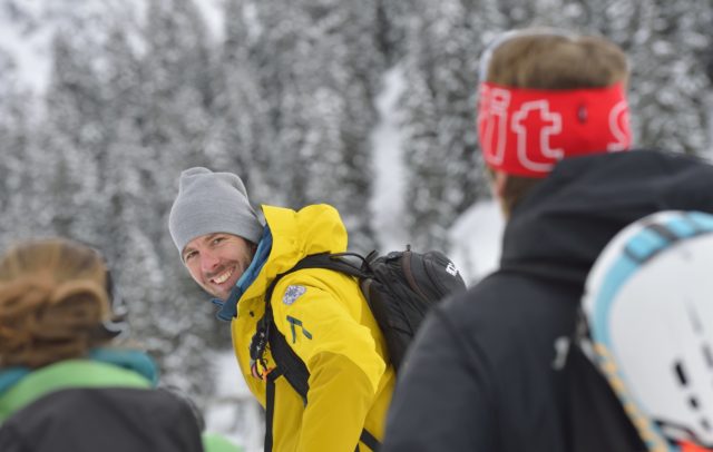 Ein Bergführer in Winterkleidung dreht sich lachend zu seinem Gast um, der nur von hinten zu sehen ist.