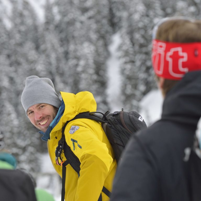Ein Bergführer in Winterkleidung dreht sich lachend zu seinem Gast um, der nur von hinten zu sehen ist.