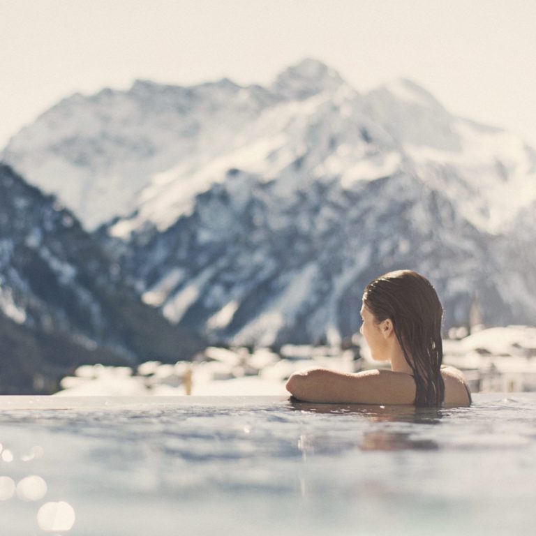 Eine Frau im Outdoorpool im Winter, im Hintergrund verschneite Berglandschaft im Kleinwalsertal, Wellness im Kleinwalsertal