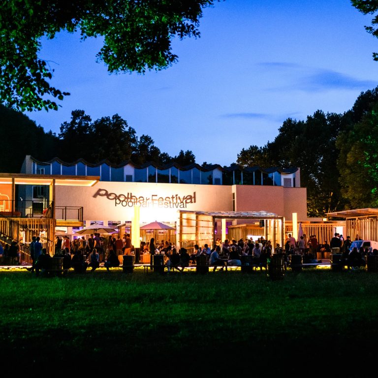Eine Aufführung des poolbar festivals, das Pubilkum sitzt im Freien vor der Bühne, schöne Abendstimmung