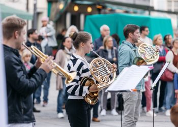Musikerinnen mit Blechblasinstrumenten spielen im Freien, Zuhörer:innen rundherum bei OUTREACH, einem, neuen Konzertformat des Symphonieorchester Vorarlberg