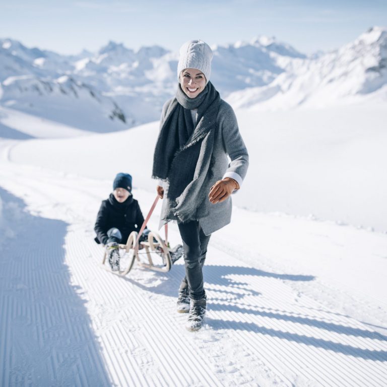 Winterwandern - eine lachende Frau auf einem verschneiten Winterwanderweg am Arlberg zieht ein Kind auf dem Rodel hinter sich her