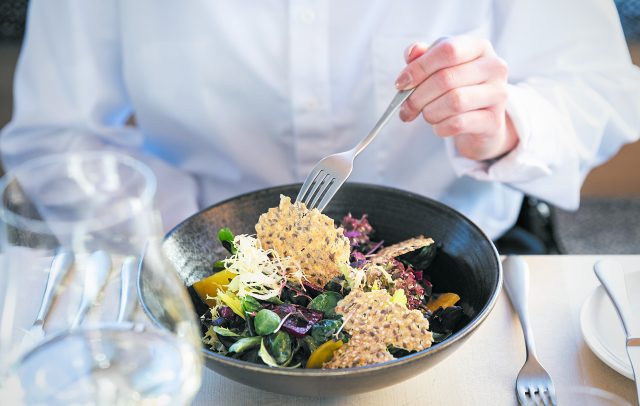 Ein gemischter Salat, serviert auf einem schwarzen Teller steht auf einem Tisch, dahinter eine Person in einer weißen Bluse, die mit der Gabel in den Salat sticht