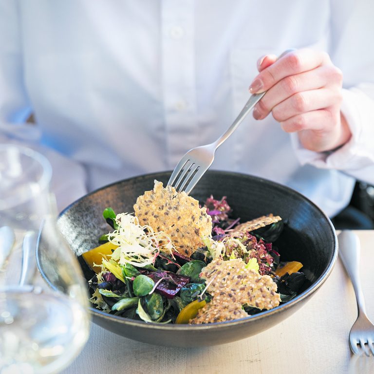 Ein gemischter Salat, serviert auf einem schwarzen Teller steht auf einem Tisch, dahinter eine Person in einer weißen Bluse, die mit der Gabel in den Salat sticht