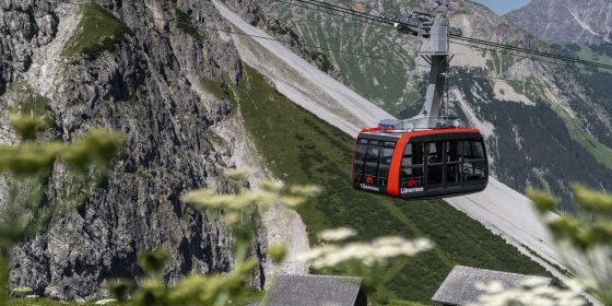 Rote Lünerseebahn-Gondel vor einer imposanten alpinen Berglandschaft mit felsigen Hängen, grünen Wiesen und blauem Himmel.