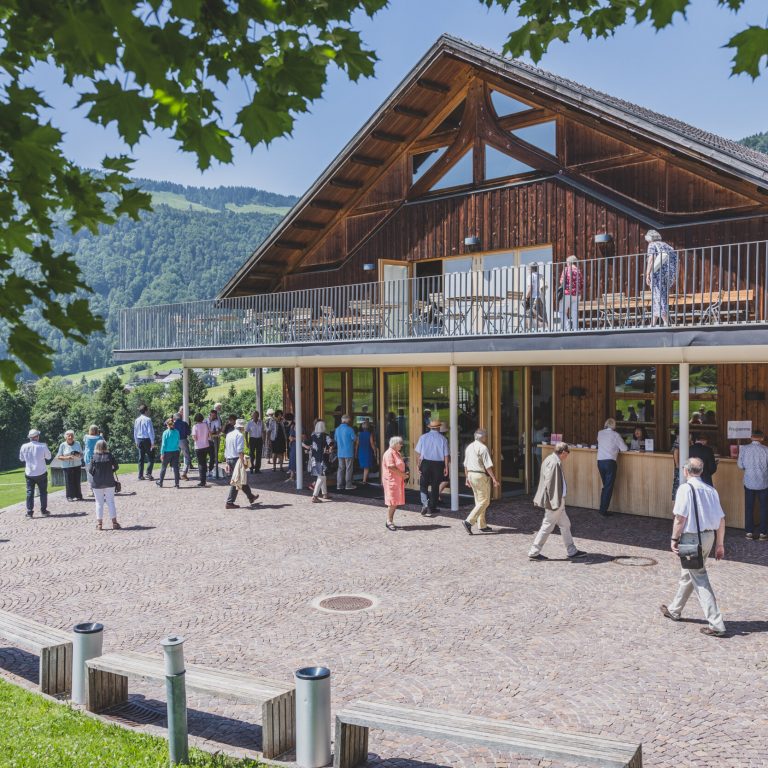 Der Angelika-Kauffmann-Saal in Schwarzenberg öffnet für die Schubertiade im Juni seine Türen. Menschen auf dem Vorplatz, die zu einem Konzert eintreffen. Im Hintergrund die grüne Landschaft des Bregenzerwaldes.