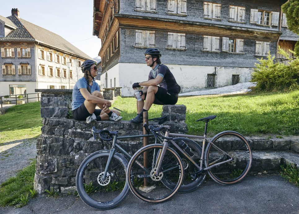 Zwei Gravelbiker in Schwarzenberg bei einer Pause, im Hintergrund zwei typische Bregenzerwälder Häuser