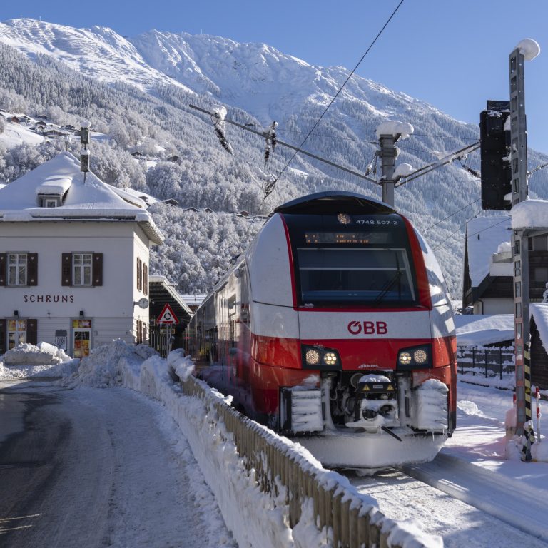 Ein Zug der ÖBB steht im Bahnhof Schruns. Das quadratische, weißliche Bahnhofsgebäude ist links im Bild ebenfalls zu sehen. All das bei Sonnenschein in winterlicher, verschneiter Berglandschaft