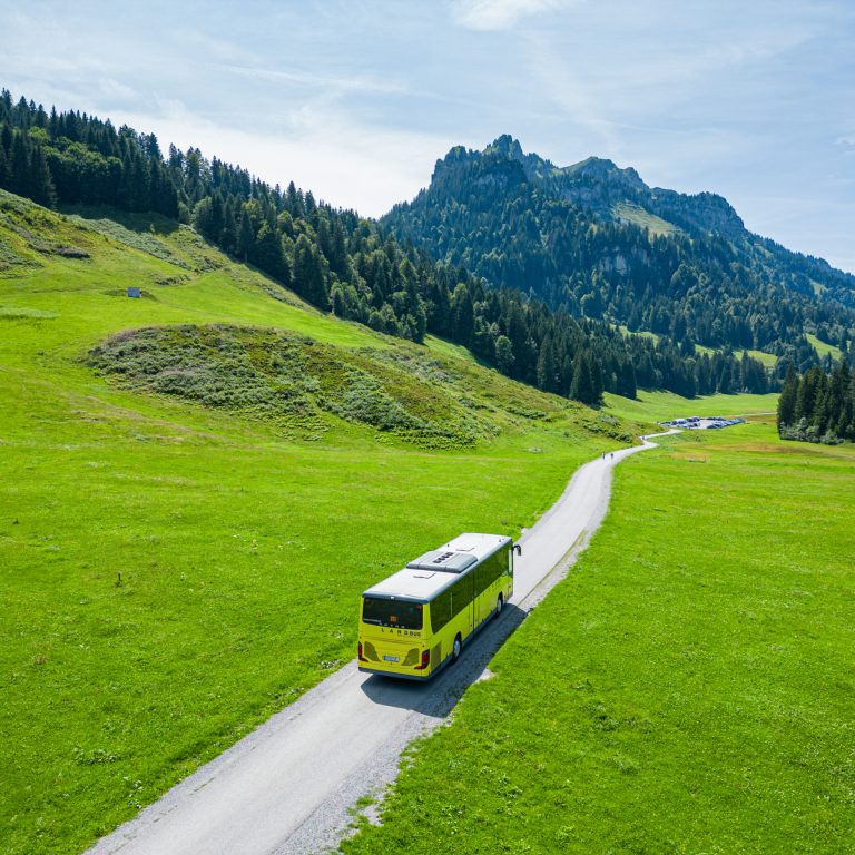 A country bus on the idyllic road to Schönenbach in the Bregenzerwald, surrounded by green fields and wooded hills.