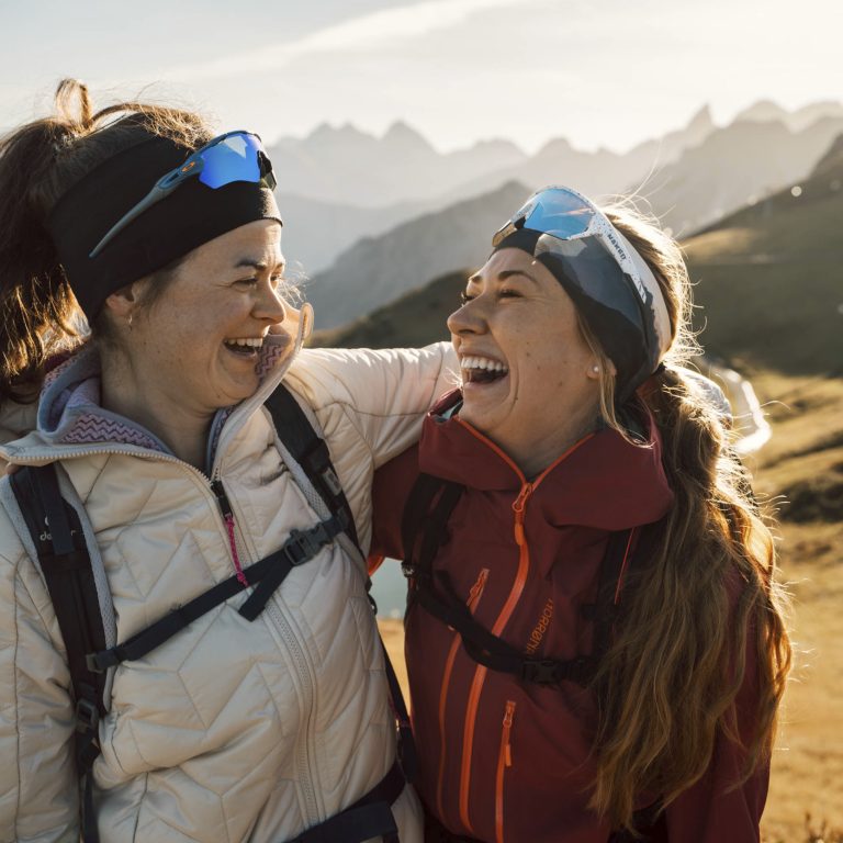 Zwei Frauen in sportlicher Kleidung schauen sich an, sie sind fröhlich. Im Hintergrund eine herbstliche Landschaft mit Blick auf Berggipfel.