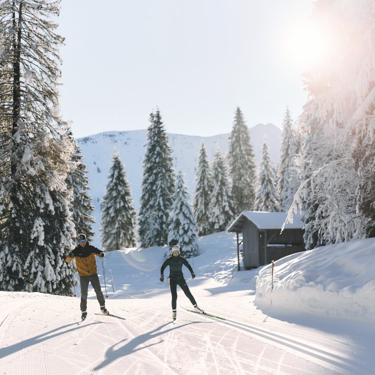 Zwei Langläufer skaten auf einer Langlaufloipe im Kleinwalsertal mit Langlaufski und Langlaufstöcke durch die verschneite Winterlandschaft.