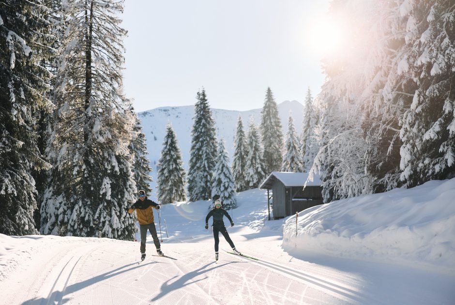 Zwei Langläufer skaten auf einer Langlaufloipe im Kleinwalsertal mit Langlaufski und Langlaufstöcke durch die verschneite Winterlandschaft.