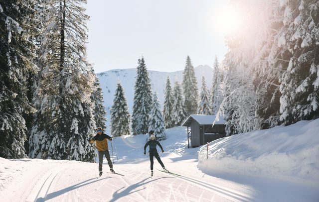 Zwei Langläufer skaten auf einer Langlaufloipe im Kleinwalsertal mit Langlaufski und Langlaufstöcke durch die verschneite Winterlandschaft.