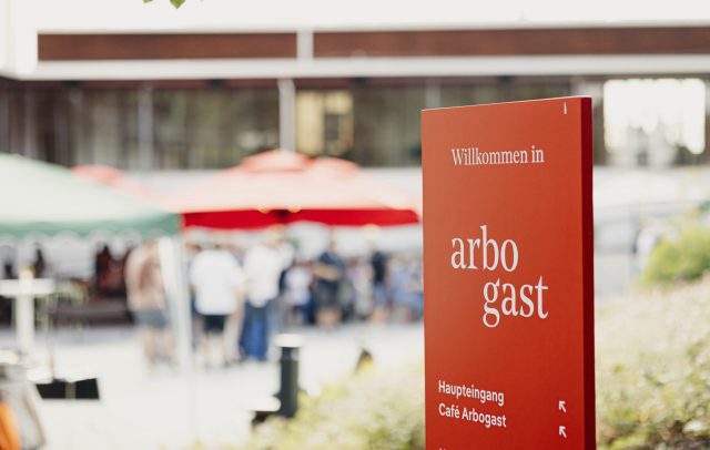 Begrüßungsschild mit der Aufschrift arbogast, im Hintergrund verschwommen Menschen im Gespräch im Freien