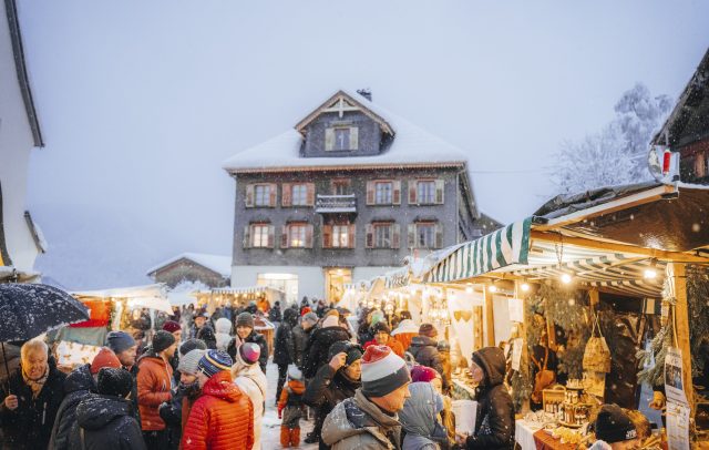 Adventmarkt in Schwarzenberg im Bregenzerwald, Menschen vor den beleuchteten Marktständen, im Hintergrund ein typisches Bregenzerwälder Haus