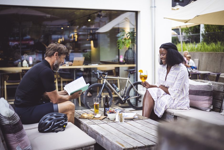 Eine Frau und ein Mann sitzen im Gastgarten eines Cafés, der Mann ist sportlich gekleidet, die Frau hat ein Kleid an, der Mann liest in der Speisekarte