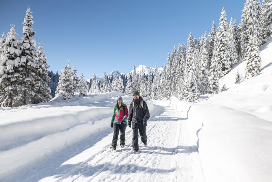 Ein Paar wandert auf einem präparierten Weg durch die verschneite Landschaft in Zug am Arlberg, im Hintergrund tief verschneite Tannen; Sanfter Winter abseits der Pisten in Vorarlberg