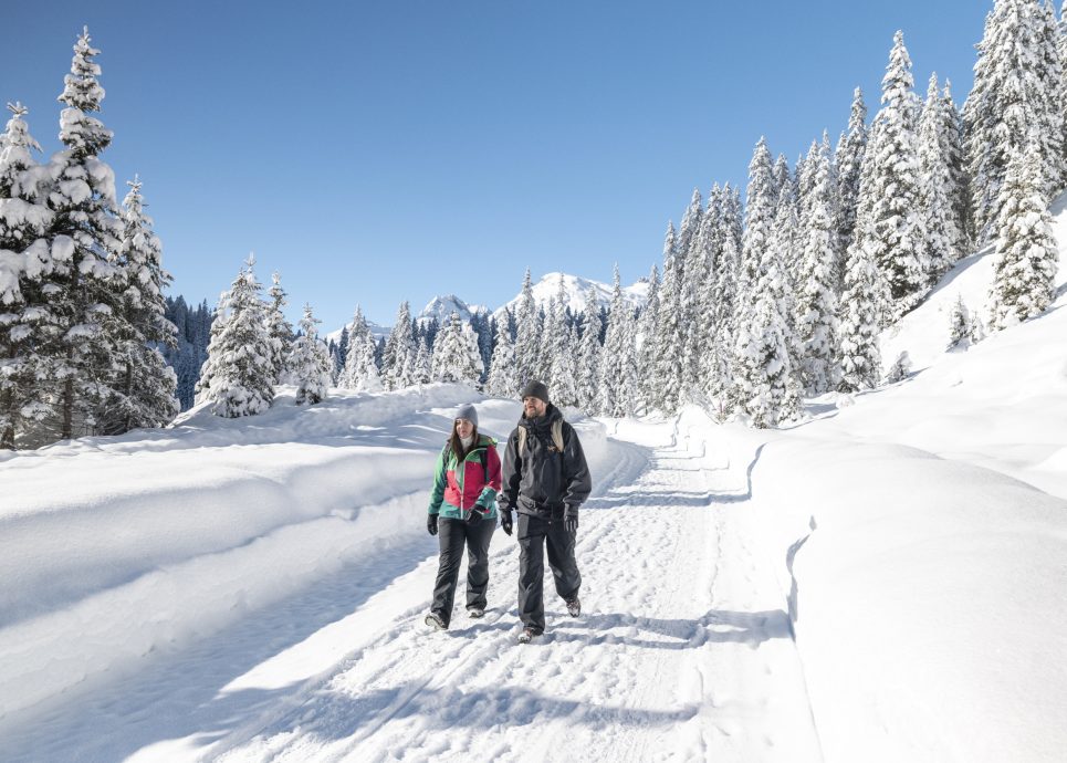 Ein Paar wandert auf einem präparierten Weg durch die verschneite Landschaft in Zug am Arlberg, im Hintergrund tief verschneite Tannen; Sanfter Winter abseits der Pisten in Vorarlberg