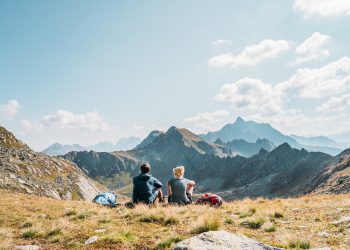 Zwei Wanderer, ein Mann und eine Frau, genießen sitzend den weiten Ausblick in die Berge beim Alpgues Rundweg im herbstlichen Montafon