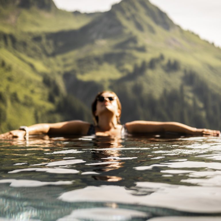 Eine Frau entspannt während ihres Wellnessurlaubs im Pool, im Hintergrund sind die Bregenzerwälder Berge zu sehen.