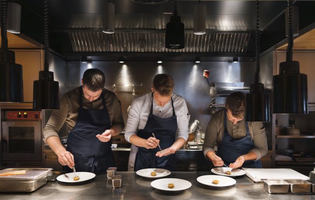 Der Sternekoch Julian Stieger vom Chef's Table Rote Wand richtet mit zwei weiteren Köchen eine Speise auf einer silbernen Arbeitsplatte an.