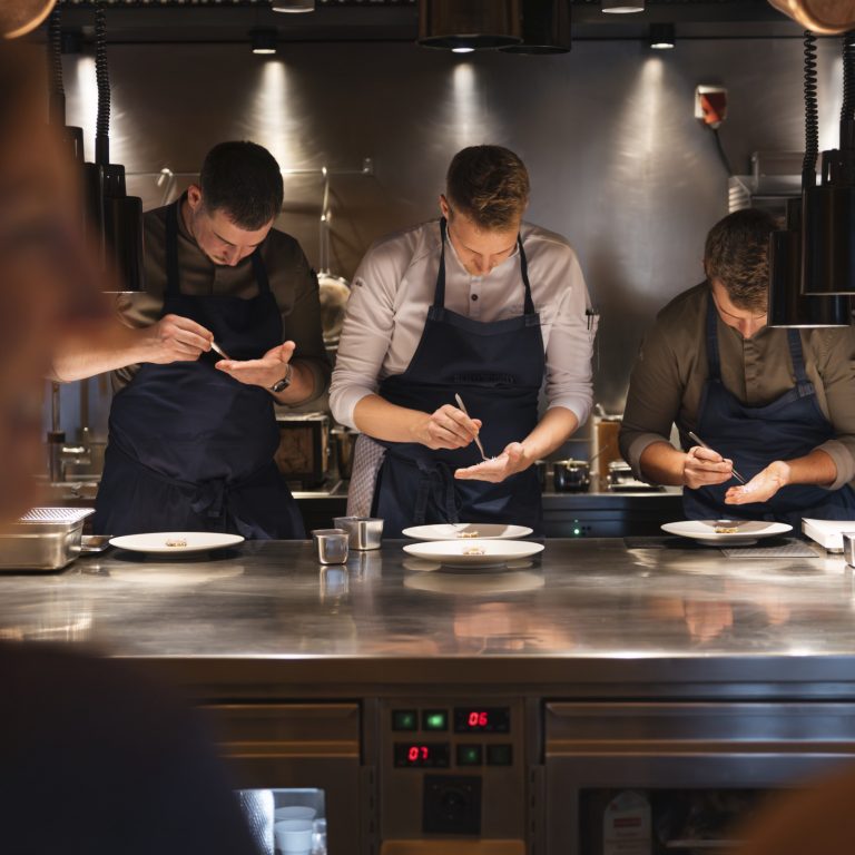 Der Sternekoch Julian Stieger vom Chef's Table Rote Wand richtet mit zwei weiteren Köchen eine Speise auf einer silbernen Arbeitsplatte an.