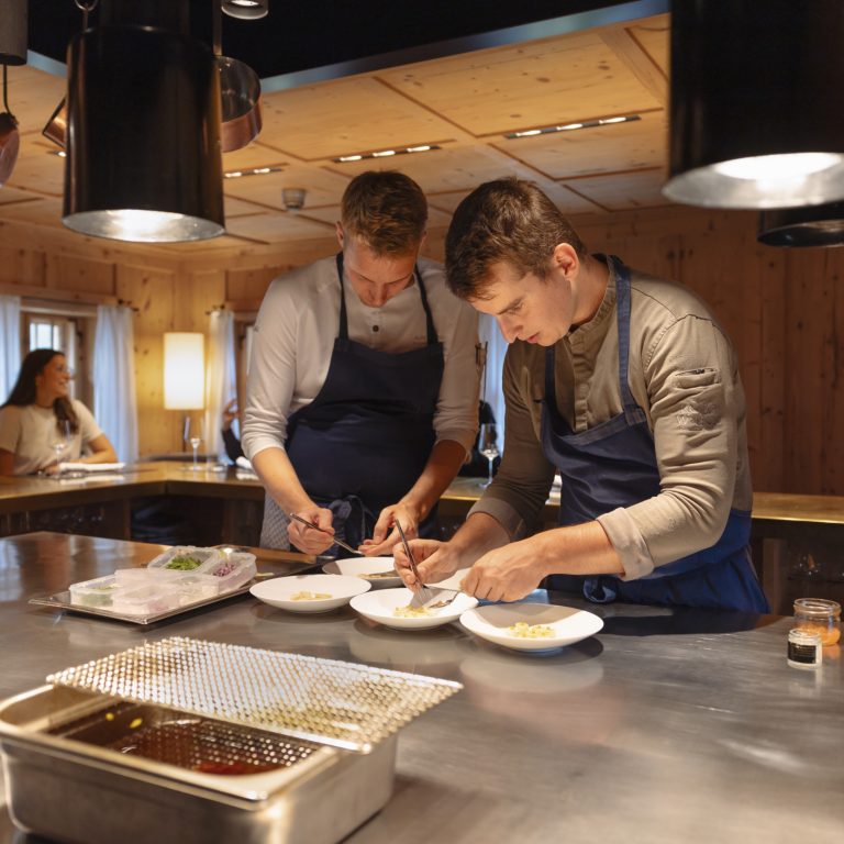 Der Sternekoch Julian Stieger vom Chef's Table Rote Wand richtet mit einem weiteren Koch eine Speise auf einer silbernen Arbeitsplatte an.