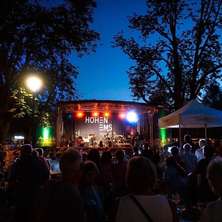 Hohenems Jazzt, ein Abed mit Konzertbesuchern, die auf Bänken sitzen und der Bühne mit Musikern im Hintergrund am Abend, Veranstaltungen in Vorarlberg