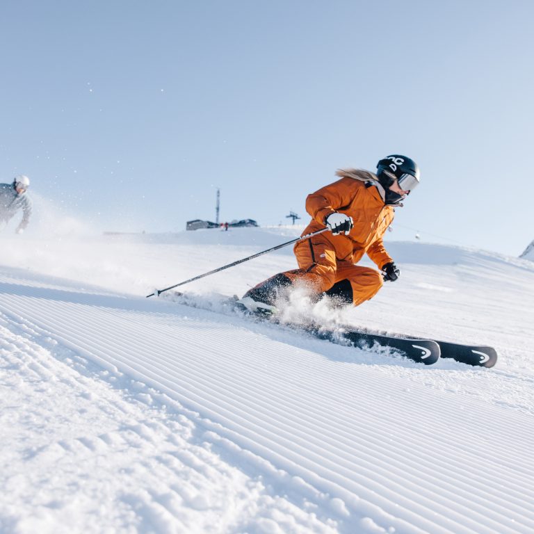 Eine weibliche Person in einem orangen Skianzug fährt mit Skiern die Piste hinunter, im Hintergrund blauer Himmel.