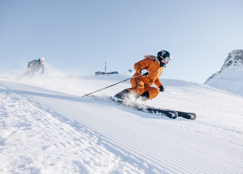 Eine weibliche Person in einem orangen Skianzug fährt mit Skiern die Piste hinunter, im Hintergrund blauer Himmel.