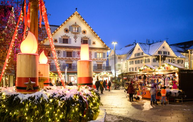 Christkindlmarkt am Marktplatz in Dornbirn, im Vordergrund ein großer Adventkrant, im Hintergrund Besucher:innen an beleuchteten Ständen