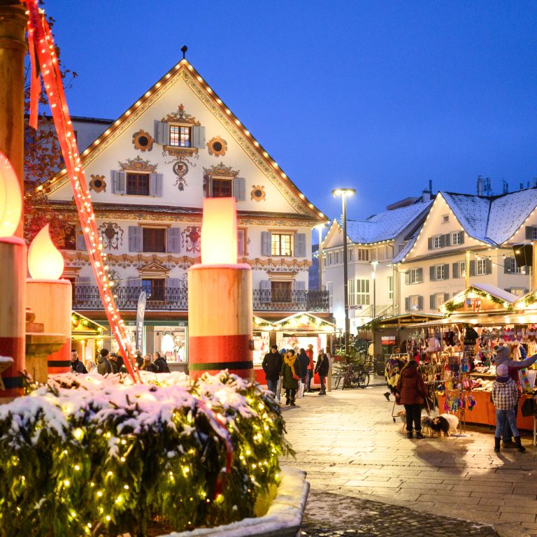 Christkindlmarkt am Marktplatz in Dornbirn, im Vordergrund ein großer Adventkrant, im Hintergrund Besucher:innen an beleuchteten Ständen