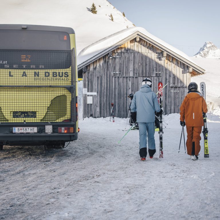 Ein gelber Landbus auf verschneiter Straße in einem Skigebiet im Bregenzerwald, Menschen mit Skiern sind gerade ausgestiegen