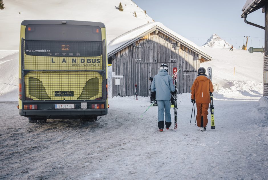 Ein gelber Landbus auf verschneiter Straße in einem Skigebiet im Bregenzerwald, Menschen mit Skiern sind gerade ausgestiegen