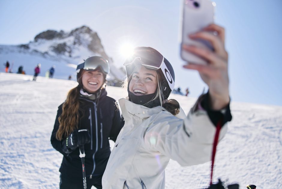 Zwei Mädchen in Skikleidung mit Helm beim Selfie-Shot, im Hintergrund Kanzelwand im Kleinwalsertal