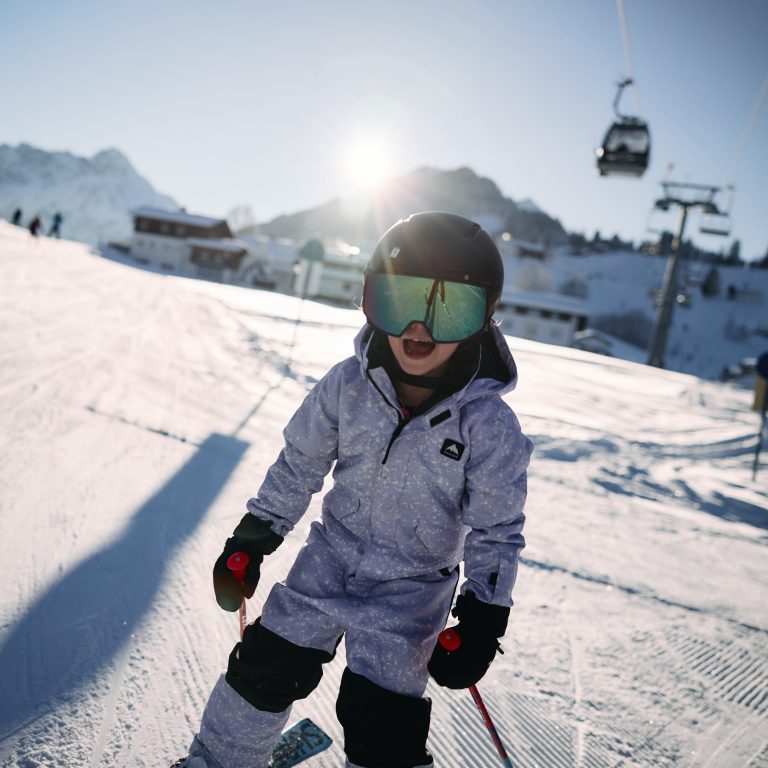 Ein Kind in Skibekleidung auf einer Skipiste, im Hintergrund blauer Himmel, Sonnenschein und eine Gondel.