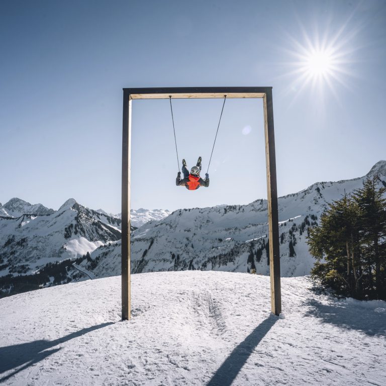 Ein Mensch auf einer großen Schaukel im verschneiten Skigebiet Damüls, die Sonne scheint vom Himmel