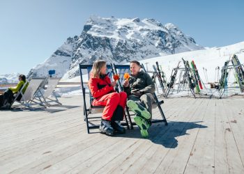 Frühlingsstimmung auf der Sonnenterrasse in Gargellen, ein Mann und eine Frau in bunter Skikleidung mit einem Getränk in der Hand, im Hintergrund schneebedeckte Berge des Montafons