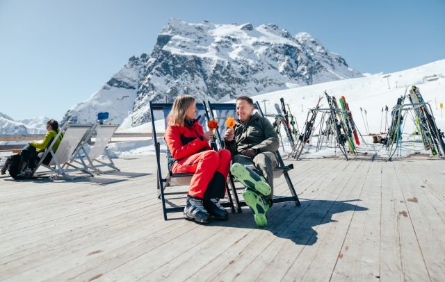 Frühlingsstimmung auf der Sonnenterrasse in Gargellen, ein Mann und eine Frau in bunter Skikleidung mit einem Getränk in der Hand, im Hintergrund schneebedeckte Berge des Montafons