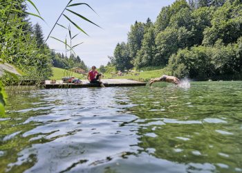 Abkühlung im Seewaldsee im Biosphärenpark Großes Walsertal, Eine Person sitzt am STeg und hält die Füße ins Wasser, Eine Person springt mit einem Kopfsprung ins Wasser.